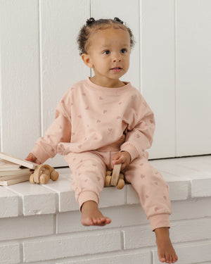 A toddler dressed in Quincy Mae’s Girls’ Relaxed Sweatpant, a cozy essential in pink covered with small heart patterns for all-day comfort, sits on a white brick ledge holding a wooden toy and gazing slightly to the side.