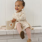 A young child wearing the Quincy Mae Baby Girls’ and Girls’ Tiered Jersey Dress sits on a white ledge, smiling and holding an open picture book, complemented by pink tights.