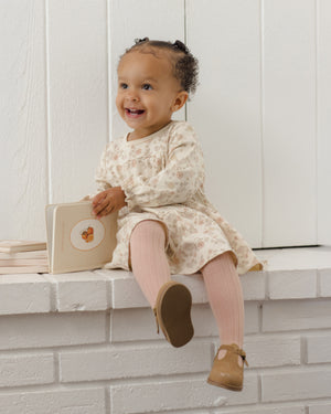 A young child wearing the Quincy Mae Baby Girls’ and Girls’ Tiered Jersey Dress sits on a white ledge, smiling and holding an open picture book, complemented by pink tights.