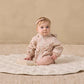 A baby, dressed in the Quincy Mae Baby and Kids’ Rain Coat featuring a pink pattern with elastic cuffs, pink tights, and a headband, sits on a round quilted mat atop a textured beige rug against a plain beige wall.