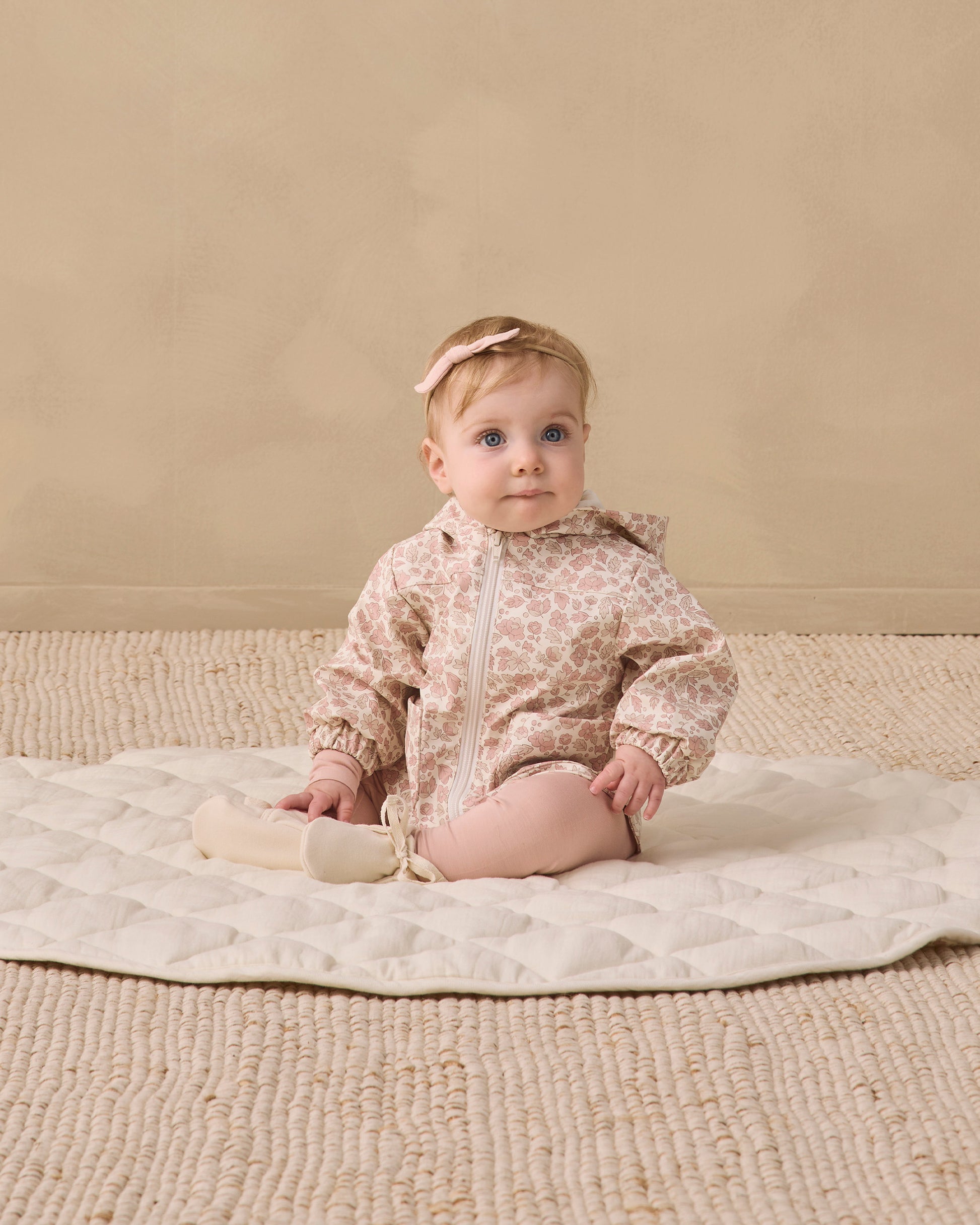 A baby, dressed in the Quincy Mae Baby and Kids’ Rain Coat featuring a pink pattern with elastic cuffs, pink tights, and a headband, sits on a round quilted mat atop a textured beige rug against a plain beige wall.