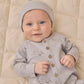 A baby wears the Quincy Mae Baby Boys’ Long Sleeve Jumpsuit in light grey, featuring coconut buttons and inseam snaps, while lying on a beige quilted blanket with a slight smile and one hand raised.