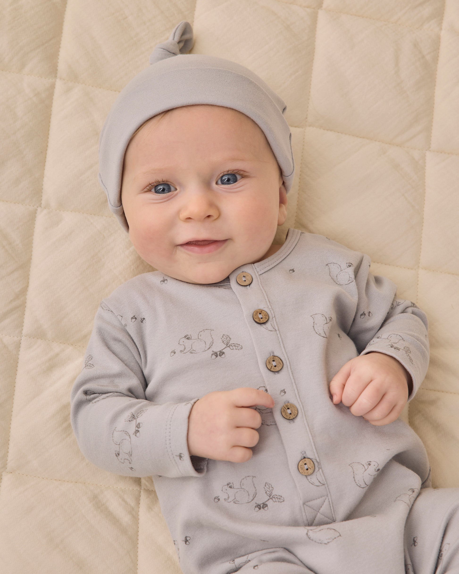 A baby wears the Quincy Mae Baby Boys’ Long Sleeve Jumpsuit in light grey, featuring coconut buttons and inseam snaps, while lying on a beige quilted blanket with a slight smile and one hand raised.