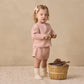 A young child dressed in the Quincy Mae Baby Girls’ and Girls’ Flynn Set in pink waffle knit with socks stands on a textured rug, holding pinecones beside a wooden basket filled with pinecones and a wooden toy, set against a beige background—perfect for everyday wear.