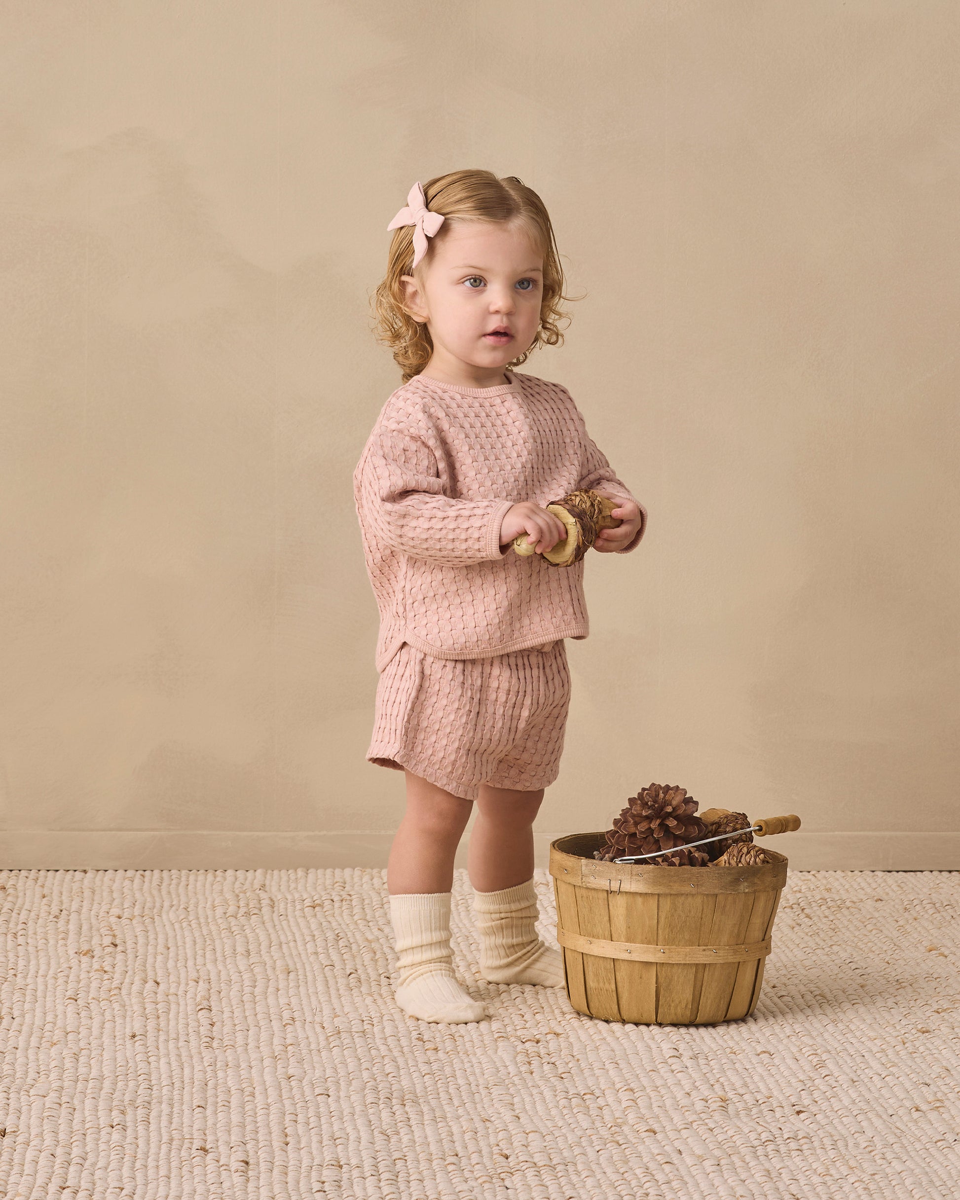 A young child dressed in the Quincy Mae Baby Girls’ and Girls’ Flynn Set in pink waffle knit with socks stands on a textured rug, holding pinecones beside a wooden basket filled with pinecones and a wooden toy, set against a beige background—perfect for everyday wear.