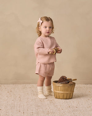 A young child dressed in the Quincy Mae Baby Girls’ and Girls’ Flynn Set in pink waffle knit with socks stands on a textured rug, holding pinecones beside a wooden basket filled with pinecones and a wooden toy, set against a beige background—perfect for everyday wear.