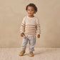 A young child with curly hair wears the Quincy Mae Baby and Kids’ Rex Sweater, showcasing cream and beige stripes with ribbed detailing. The sweater is paired with light pants and tan shoes, as the child stands on a textured rug set against a beige background.