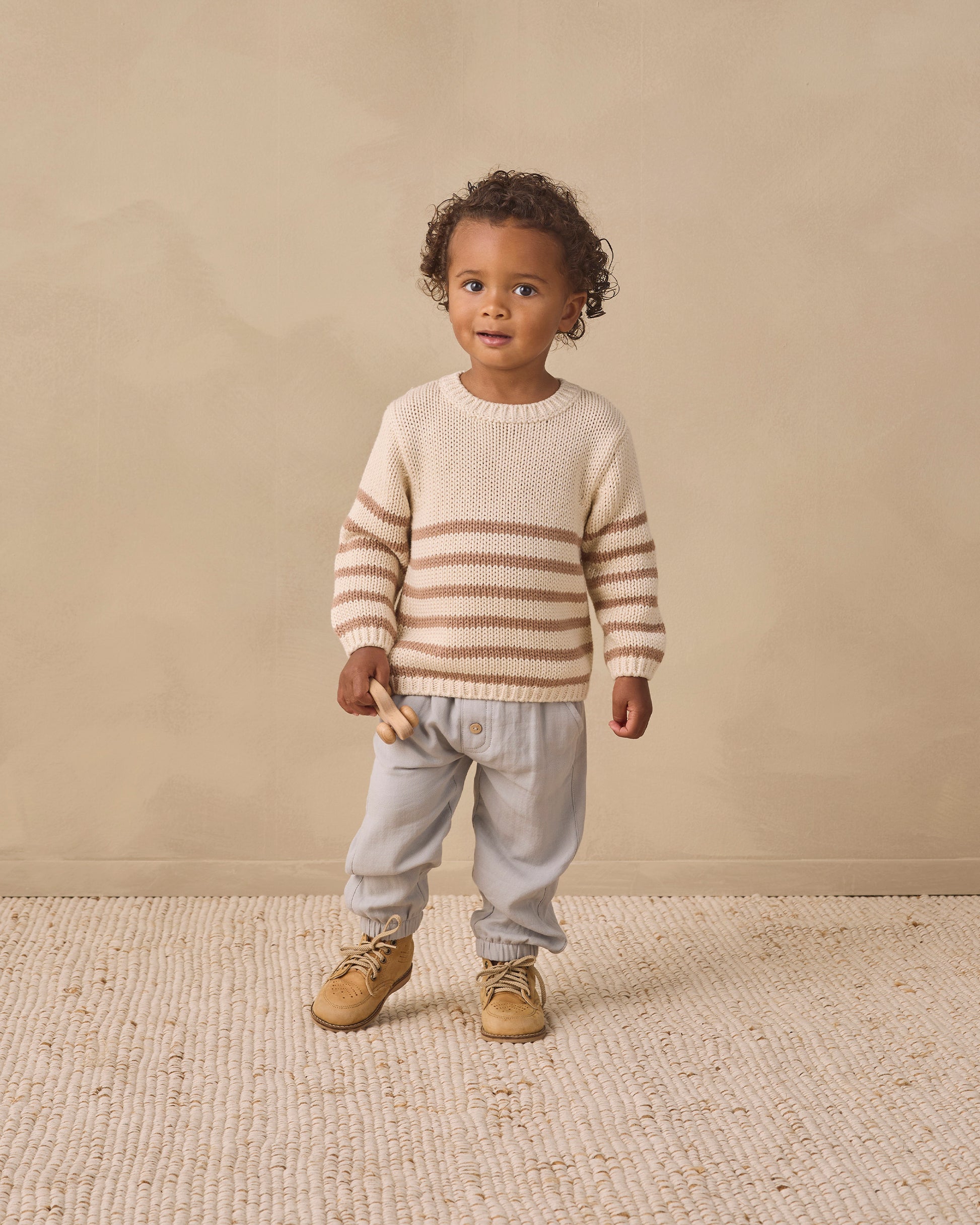 A young child with curly hair wears the Quincy Mae Baby and Kids’ Rex Sweater, showcasing cream and beige stripes with ribbed detailing. The sweater is paired with light pants and tan shoes, as the child stands on a textured rug set against a beige background.