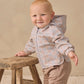 A smiling baby stands holding a wooden stool, dressed in the Quincy Mae Baby and Kids’ Rain Coat featuring a plaid hood, elastic cuffs, and paired with beige pants against a neutral background.