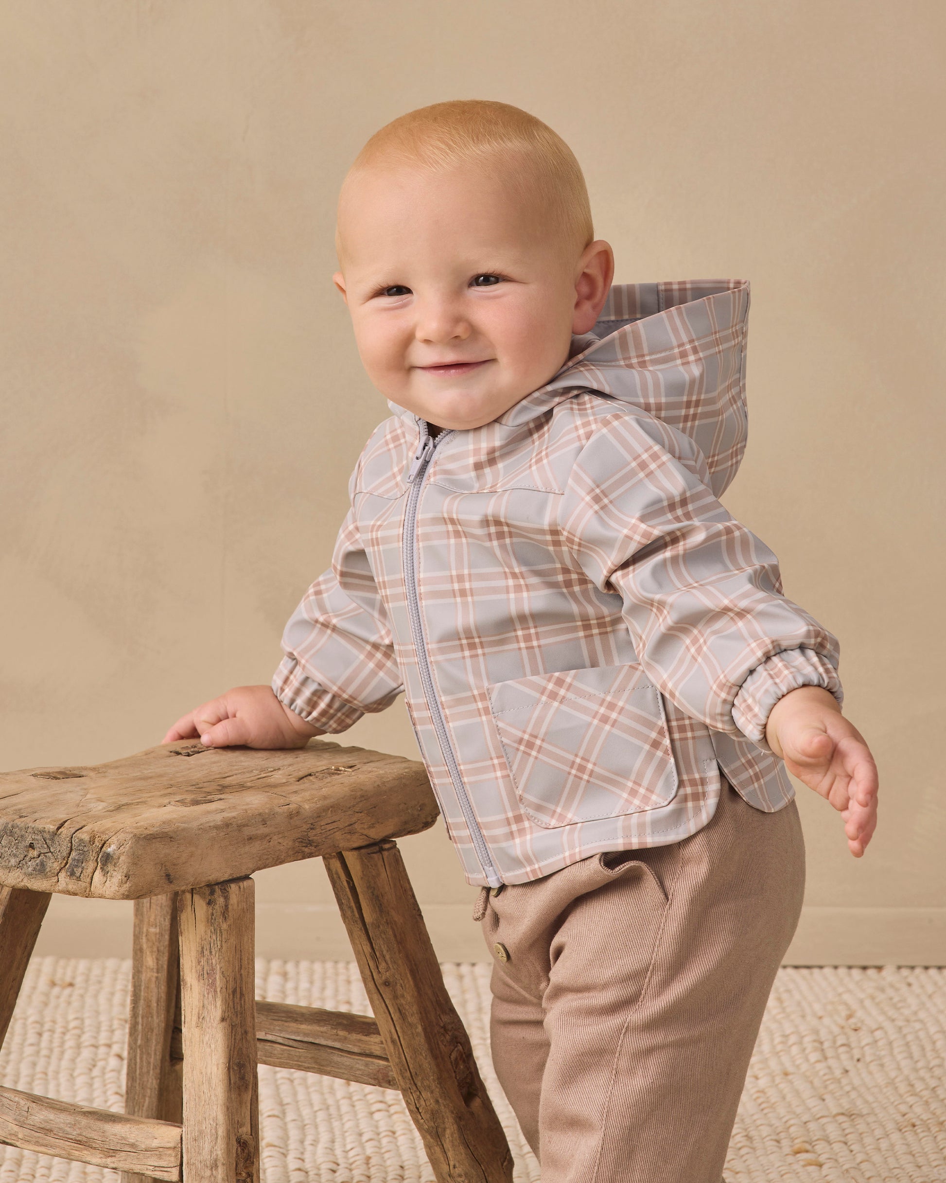 A smiling baby stands holding a wooden stool, dressed in the Quincy Mae Baby and Kids’ Rain Coat featuring a plaid hood, elastic cuffs, and paired with beige pants against a neutral background.