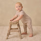 A baby wearing the Quincy Mae Baby Short Sleeve Jumpsuit in striped beige with coconut buttons stands holding a small wooden stool on a textured rug, facing slightly toward the camera—for easy changes and adorable style.