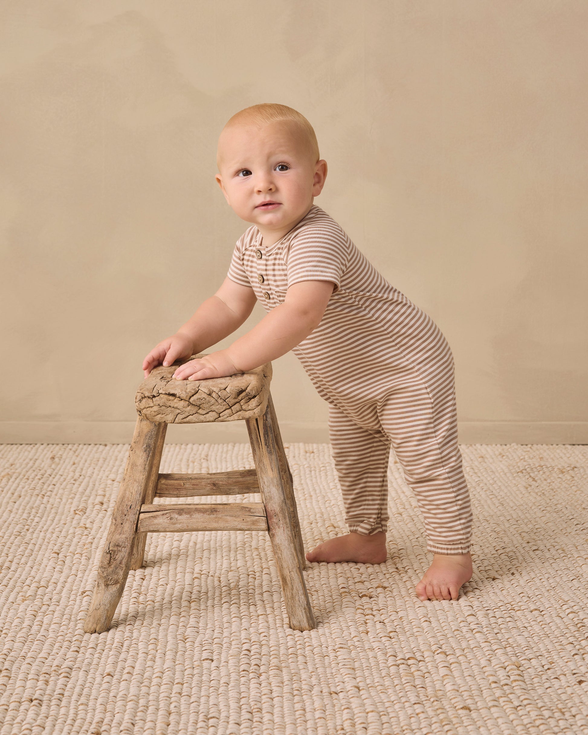 A baby wearing the Quincy Mae Baby Short Sleeve Jumpsuit in striped beige with coconut buttons stands holding a small wooden stool on a textured rug, facing slightly toward the camera—for easy changes and adorable style.