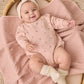 A baby lies in a wicker basket, dressed in the Quincy Mae Baby Girls’ Crewneck Bubble Romper featuring small heart patterns, paired with a headband and cream socks, resting on a textured pink blanket.