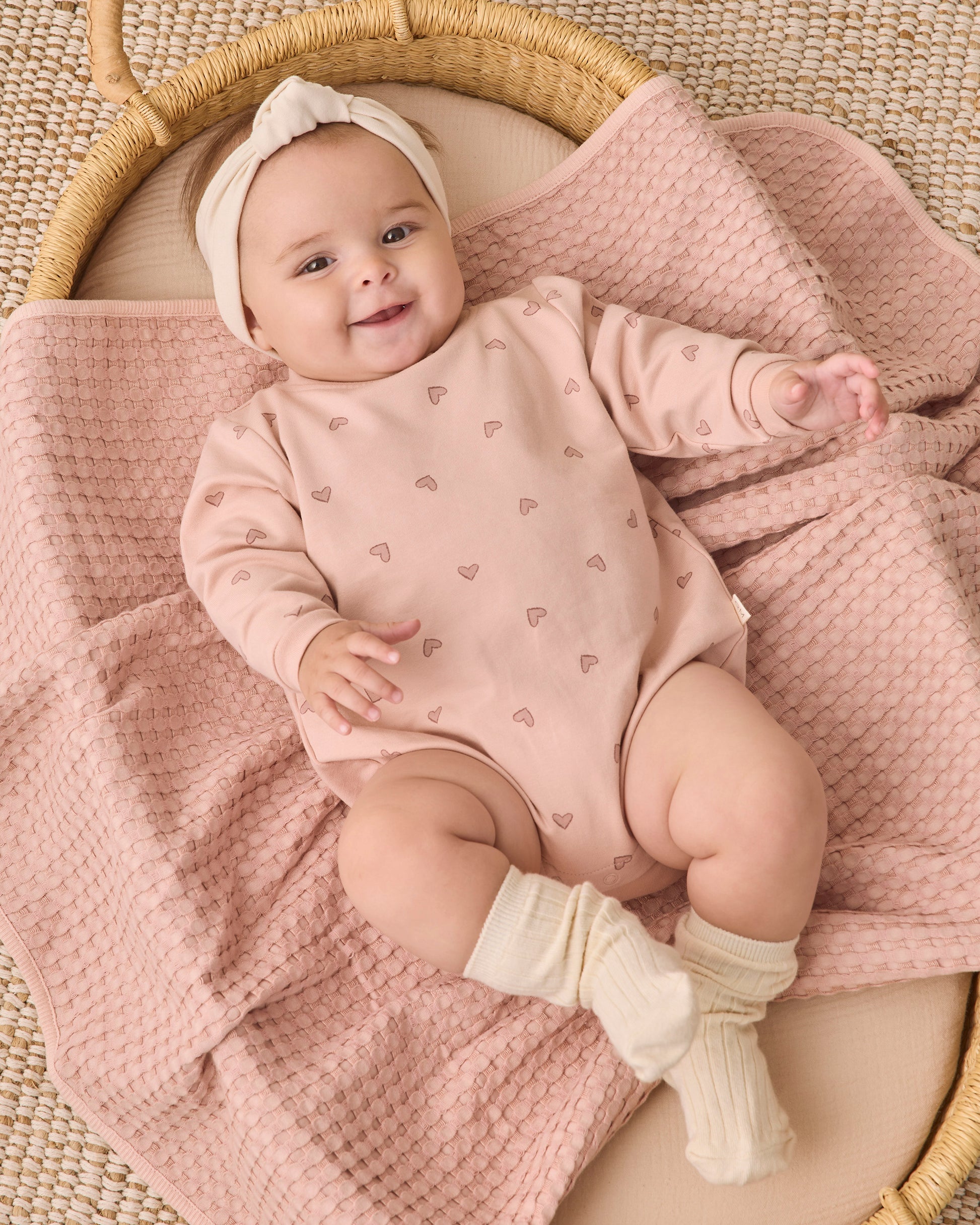 A baby lies in a wicker basket, dressed in the Quincy Mae Baby Girls’ Crewneck Bubble Romper featuring small heart patterns, paired with a headband and cream socks, resting on a textured pink blanket.
