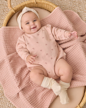 A baby lies in a wicker basket, dressed in the Quincy Mae Baby Girls’ Crewneck Bubble Romper featuring small heart patterns, paired with a headband and cream socks, resting on a textured pink blanket.