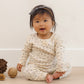 A baby sits on the floor wearing the Quincy Mae Baby Girls’ Pointelle Long Sleeve Tee + Legging set in a cream floral pattern, paired with a gray headband and surrounded by pinecones and a wooden ball, against a neutral background.