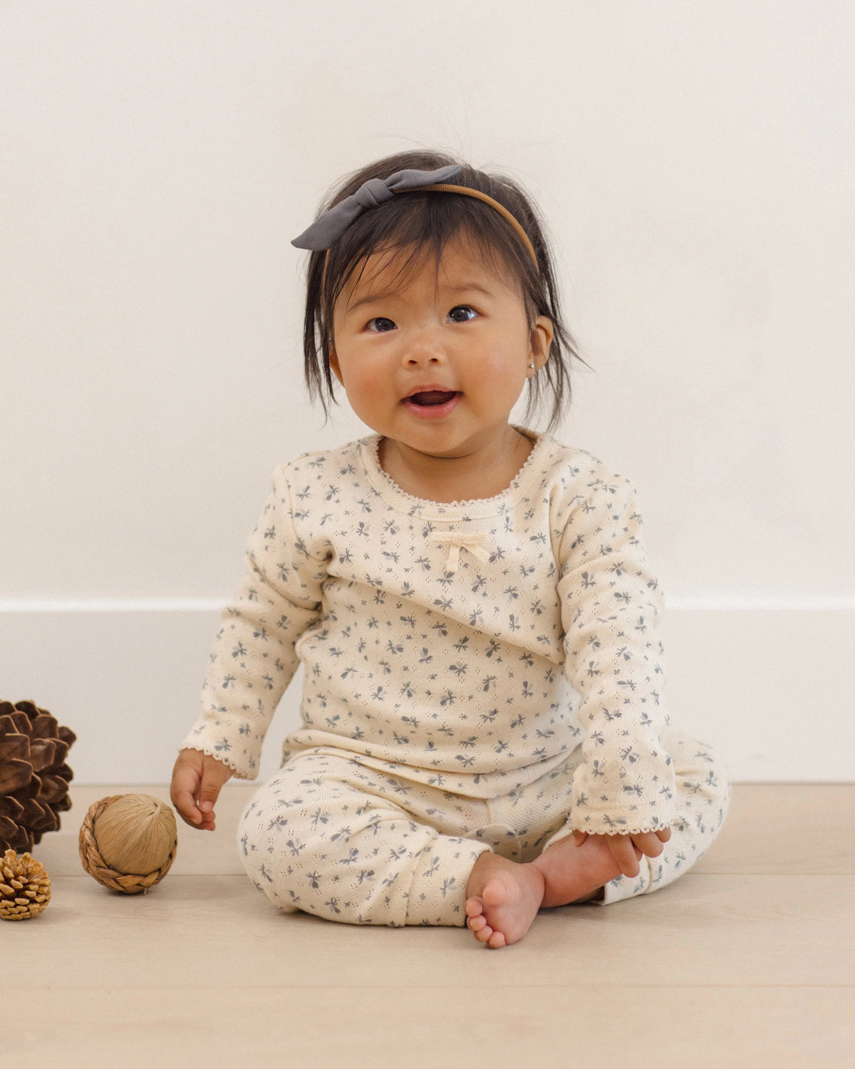 A baby sits on the floor wearing the Quincy Mae Baby Girls’ Pointelle Long Sleeve Tee + Legging set in a cream floral pattern, paired with a gray headband and surrounded by pinecones and a wooden ball, against a neutral background.