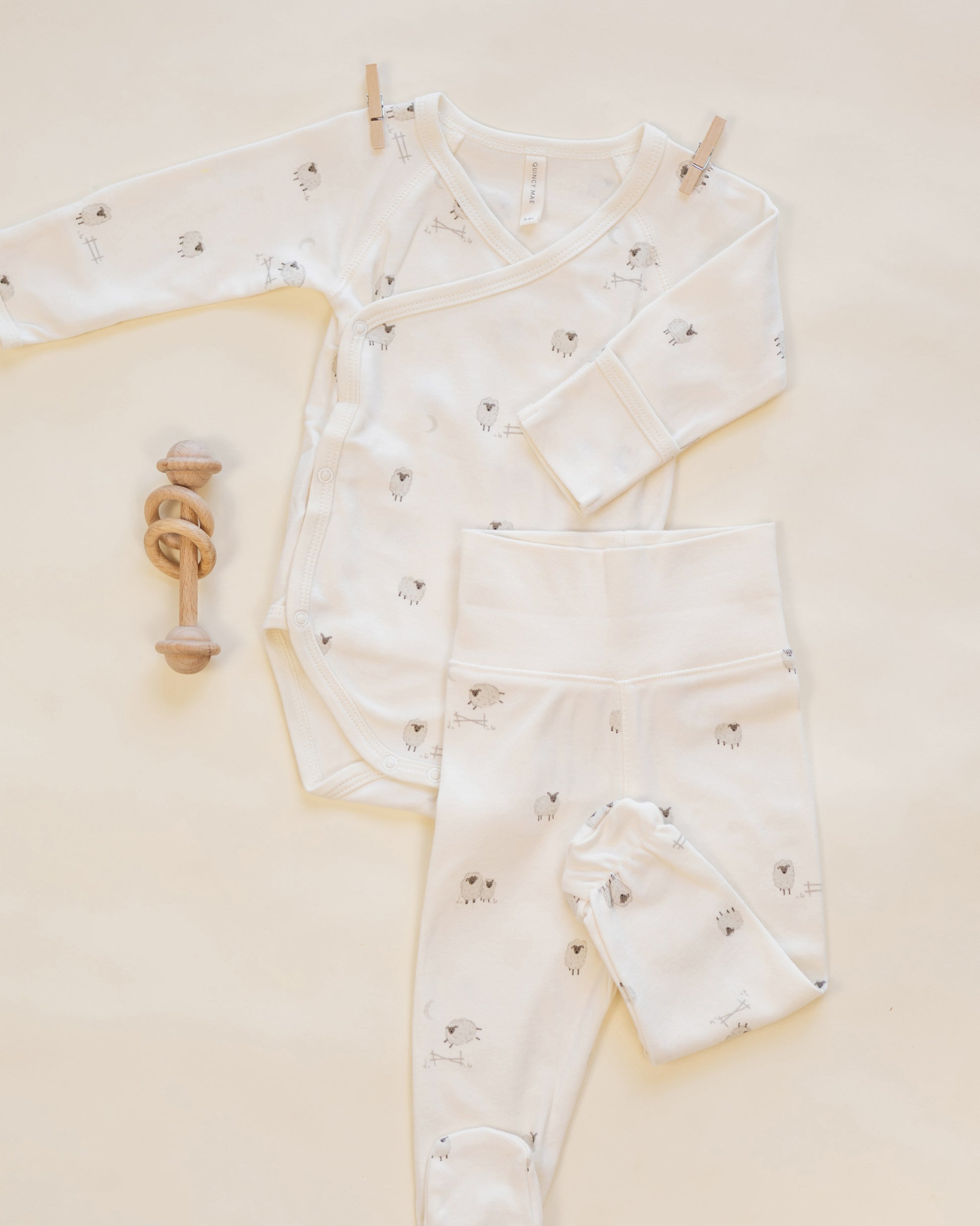 Flat lay of the Quincy Mae Baby Side Snap Bodysuit + Pant Set, featuring a white bodysuit and matching footed pants with small animal prints, displayed beside a wooden rattle on a light background—perfect for keeping your newborn cozy.