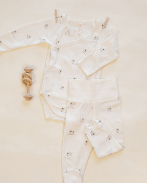 Flat lay of the Quincy Mae Baby Side Snap Bodysuit + Pant Set, featuring a white bodysuit and matching footed pants with small animal prints, displayed beside a wooden rattle on a light background—perfect for keeping your newborn cozy.