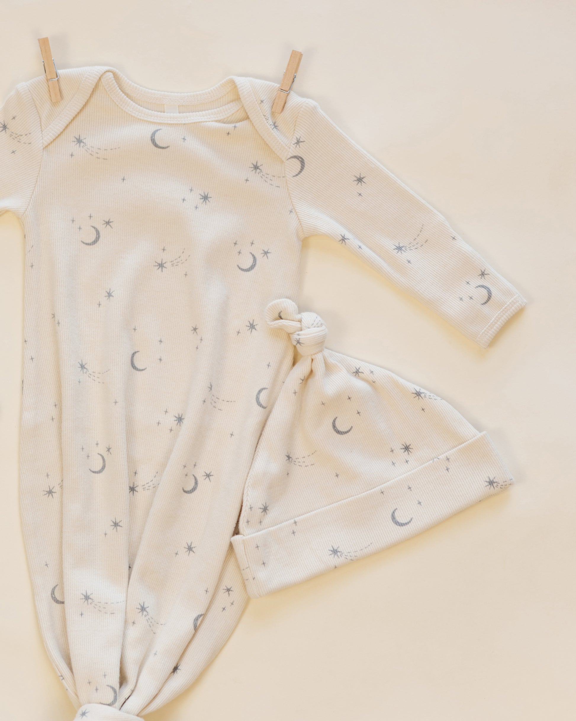 The Quincy Mae Baby Boys’ Knotted Baby Gown + Hat Set, made from soft organic rib knit and adorned with a grey moon and star pattern, is shown laid flat on a cream background with two wooden clothespins.