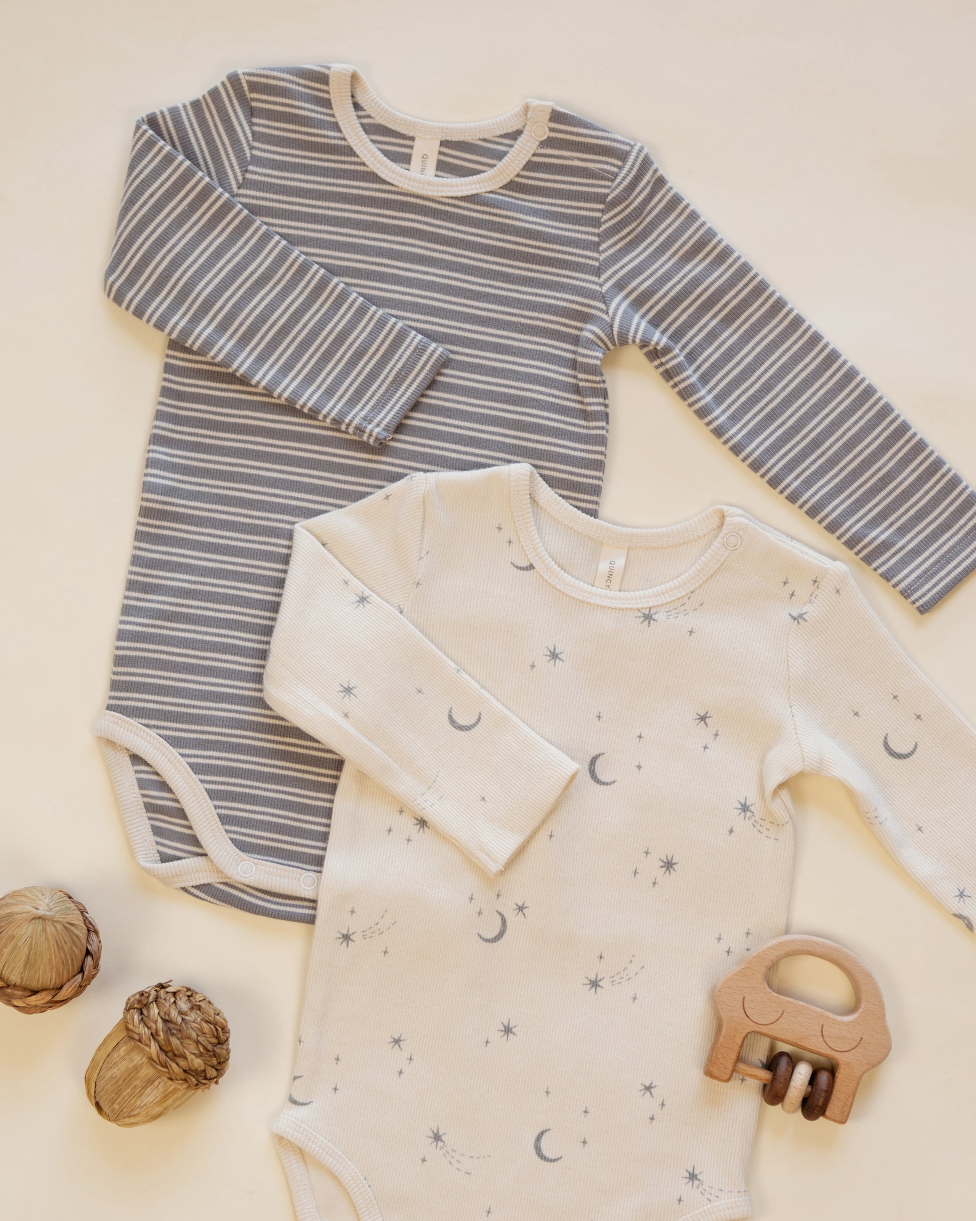 Two baby boys’ ribbed bodysuits from Quincy Mae’s 2-pack set, including one in grey with white stripes and another in cream featuring moons and stars, are displayed on a light surface alongside wooden toys and acorns.