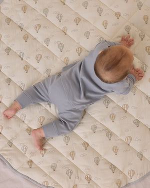A baby in a light blue Quincy Mae Baby and Kids' Long Sleeve Tee with shoulder snaps lies on their stomach on a quilted blanket featuring a hot air balloon pattern.