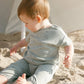 A baby wearing the Quincy Mae Baby Boys' Jersey Tee & Pant Set with dino print sits on sandy ground beneath a white umbrella, gazing downward and to the side.