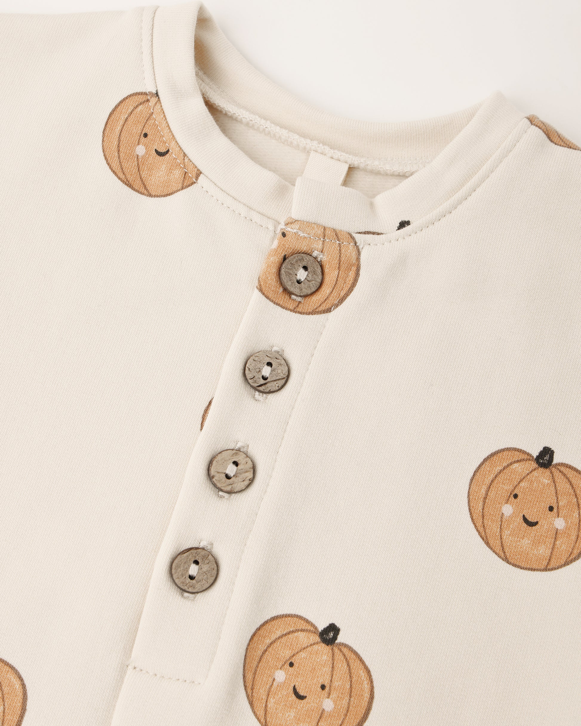 Close-up of the Quincy Mae Baby Relaxed Fleece Jumpsuit in cream, featuring three buttons and a cheerful pattern of smiling orange pumpkins—crafted for everyday comfort as a cozy essential in your little one’s wardrobe.