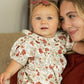 A baby in the Rylee & Cru Baby Girls' Lula Romper sits on a woman's lap, both smiling and looking the same way.