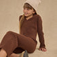 A young girl wearing a Rylee & Cru Girl's Long Sleeve Knit Button Top sits on the floor against a neutral background, smiling gently with one hand behind her and a cream knit beanie on her head.