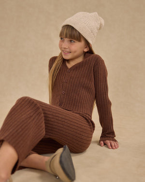 A young girl wearing a Rylee & Cru Girl's Long Sleeve Knit Button Top sits on the floor against a neutral background, smiling gently with one hand behind her and a cream knit beanie on her head.