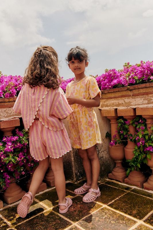 Two young girls wear the Louise Misha Baby Girls' and Girls' Chiara Playsuit on a terrace with bright pink flowers and a cloudy sky, their colorful outfits echoing playful jacquard patterns.