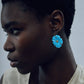 A short-haired person wearing a sleeveless beige top models Lizzie Fortunato’s New Bloom Earrings—gold-plated brass pieces shaped like blue flowers—while looking left against a plain background.