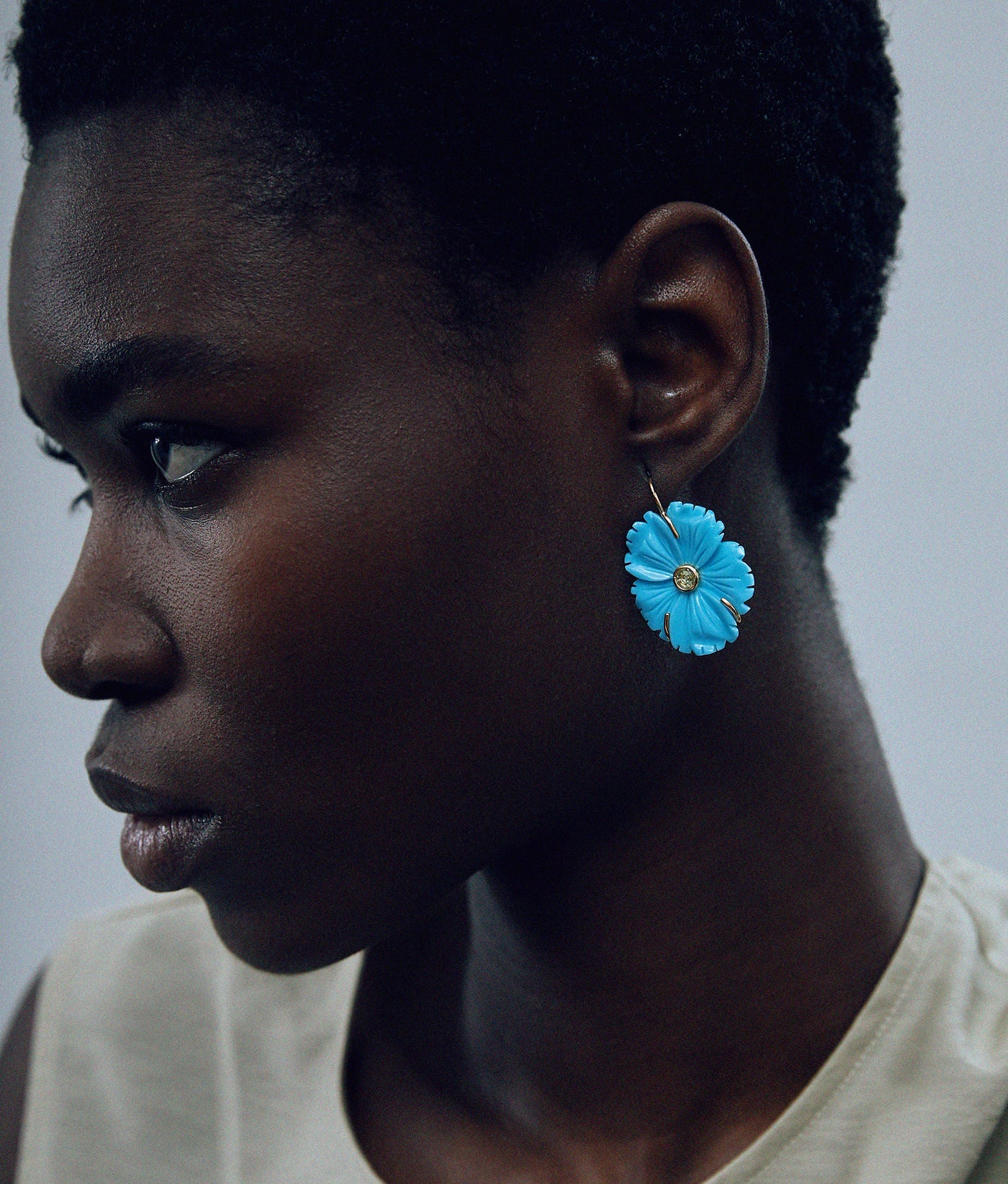 A short-haired person wearing a sleeveless beige top models Lizzie Fortunato’s New Bloom Earrings—gold-plated brass pieces shaped like blue flowers—while looking left against a plain background.