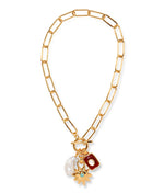 The Helios Charm Necklace by Lizzie Fortunato features a gold chain with large links, accented by a white freshwater pearl, a red rectangular charm, and a gold sunburst charm set with a small green stone.