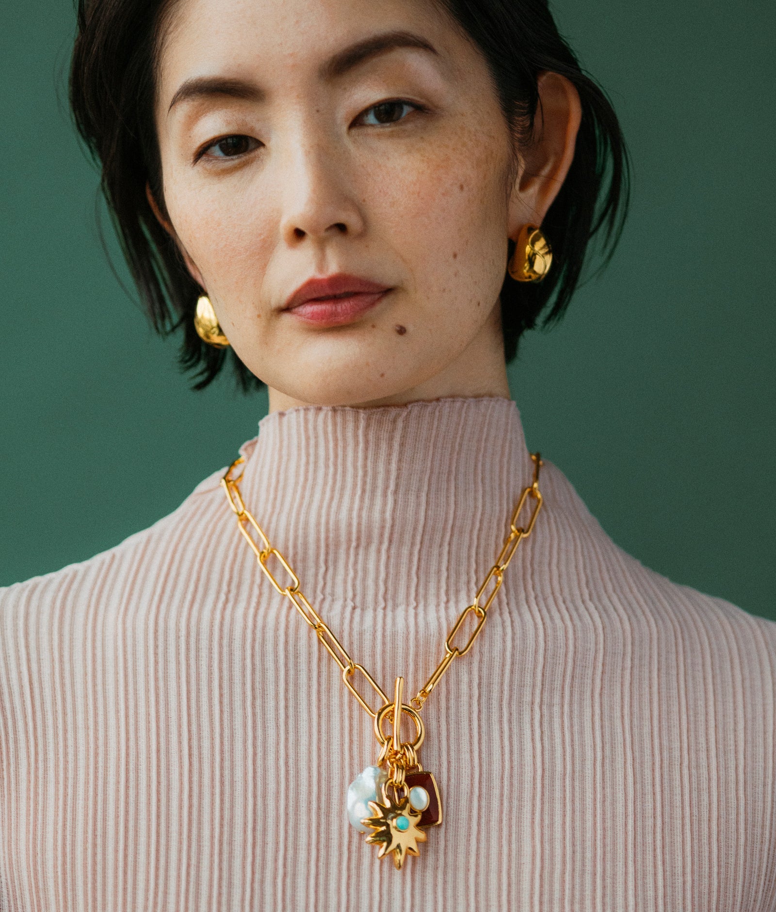 A woman wearing a ribbed, light pink turtleneck with gold earrings and the Lizzie Fortunato Helios Charm Necklace poses against a green background.