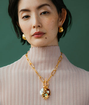 A woman wearing a ribbed, light pink turtleneck with gold earrings and the Lizzie Fortunato Helios Charm Necklace poses against a green background.