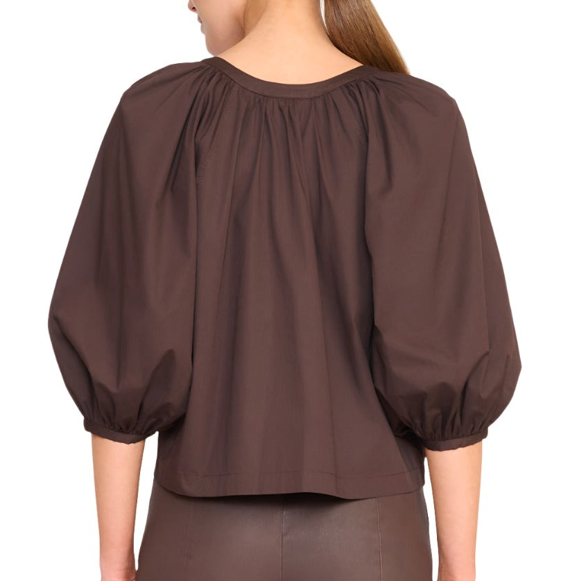 A person stands facing away, wearing a dark brown Staud Dill Top—a true closet staple—and fitted brown pants with a flared bottom.