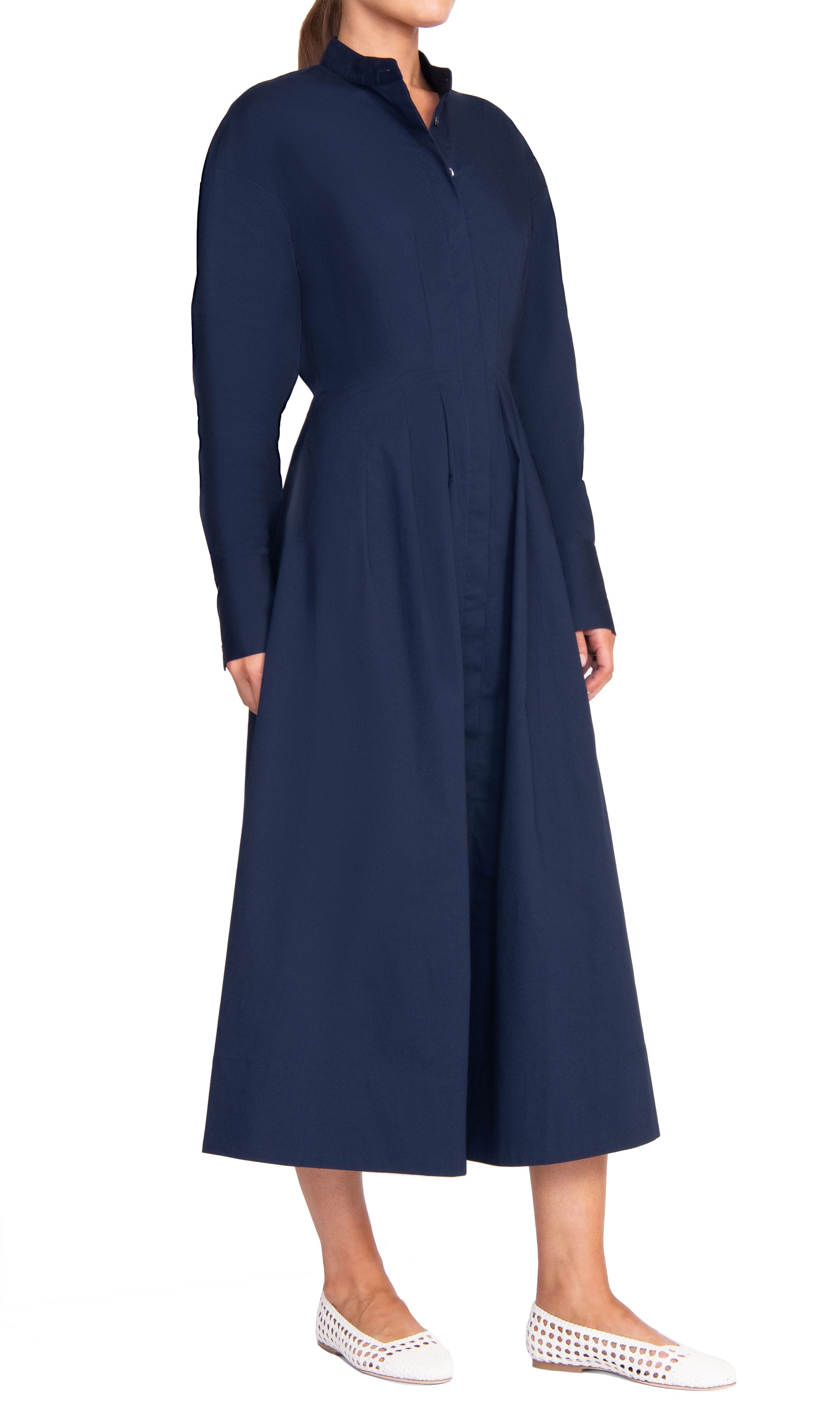 Person wearing the Staud Lorenza Midi Dress, a long button-up dress in navy blue with long sleeves and pleated details, paired with white perforated flat shoes.