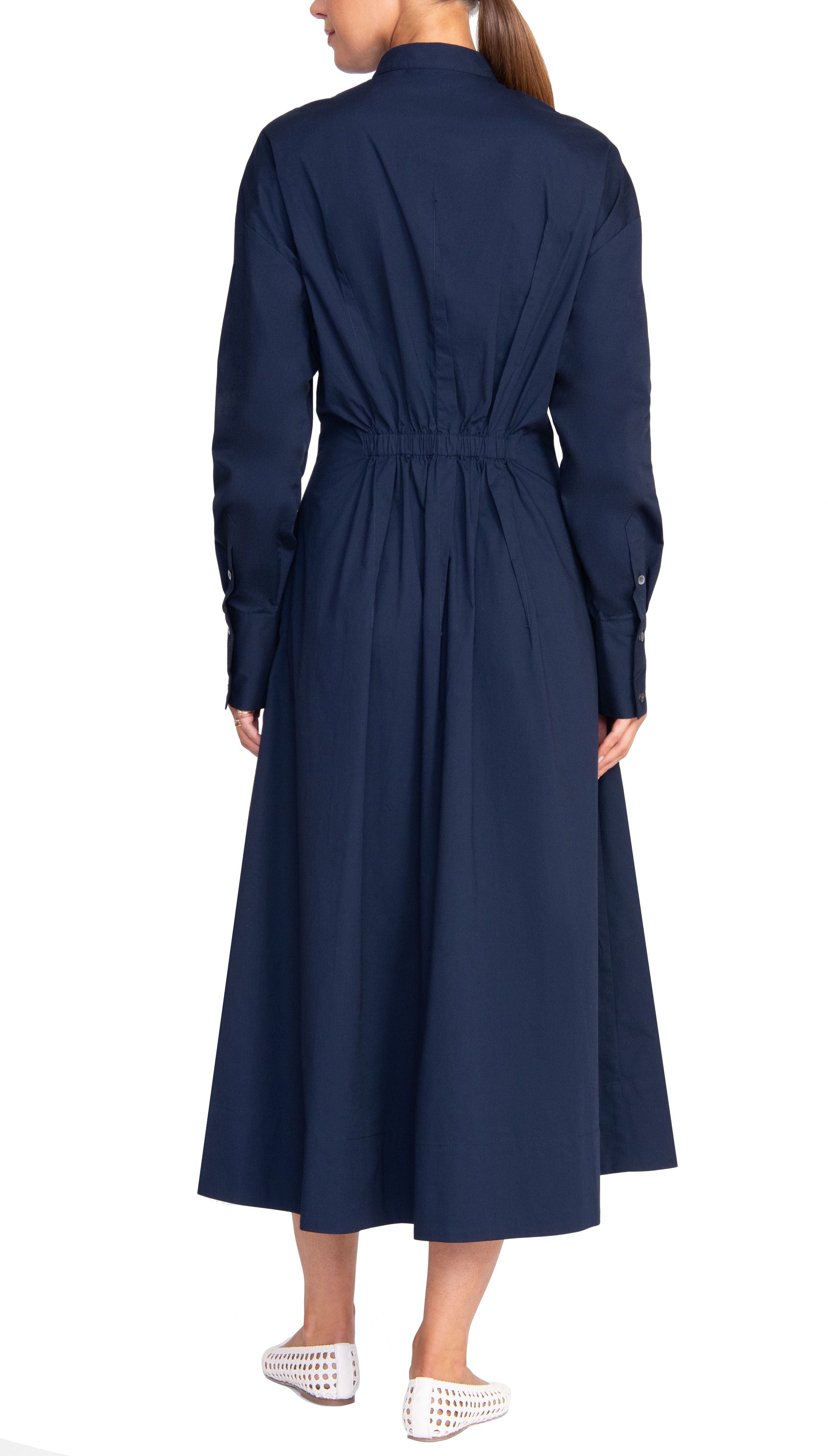 A person is shown from the back wearing the Staud Lorenza Midi Dress in navy blue, featuring long sleeves, a button-up front, and a fitted waist that reaches the calves. They are also wearing white slip-on shoes with perforations, and their hair is pulled back into a ponytail.