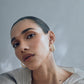 A close-up of a person with slicked-back hair wearing Lizzie Fortunato Sculpted Hoops and a pleated top, looking directly at the camera against a soft gray background.