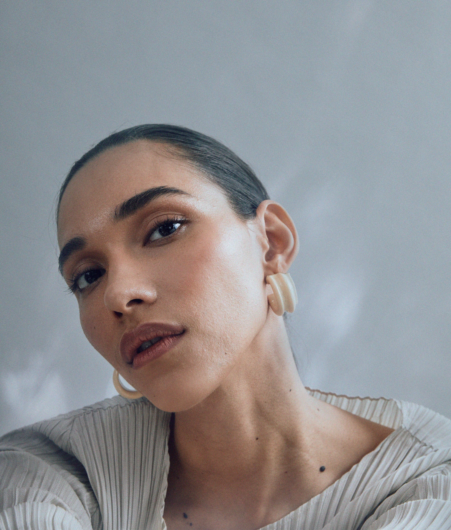 A close-up of a person with slicked-back hair wearing Lizzie Fortunato Sculpted Hoops and a pleated top, looking directly at the camera against a soft gray background.