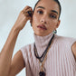Woman wearing a sleeveless, light pink ribbed turtleneck sweater and layered necklaces, accessorized with Lizzie Fortunato Organic Mini Gold Hoops and a round black ring, poses with her hand on her face against a neutral background.