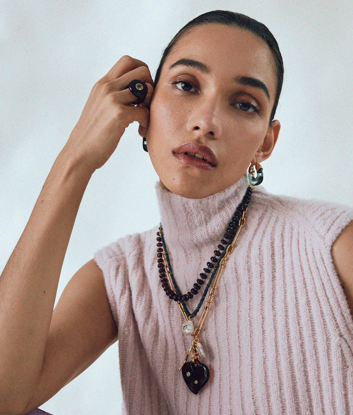 Woman wearing a sleeveless, light pink ribbed turtleneck sweater and layered necklaces, accessorized with Lizzie Fortunato Organic Mini Gold Hoops and a round black ring, poses with her hand on her face against a neutral background.