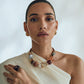 A person is wearing an off-shoulder white top, a chunky artisan-crafted beaded necklace, and the Lizzie Fortunato Halo Ring embellished with an amethyst stone, with their hair pulled back against a plain background.