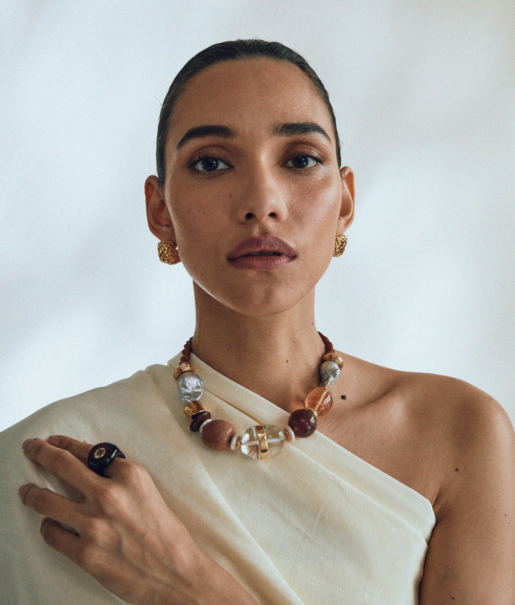 A person is wearing an off-shoulder white top, a chunky artisan-crafted beaded necklace, and the Lizzie Fortunato Halo Ring embellished with an amethyst stone, with their hair pulled back against a plain background.