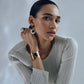 A person with slicked-back hair wears a pleated top, large earrings, and a gold bracelet, elegantly posing with their hand near their face against a neutral background. Their fingers are adorned with vibrant Lizzie Fortunato Monument Rings, each piece a wearable art masterpiece.