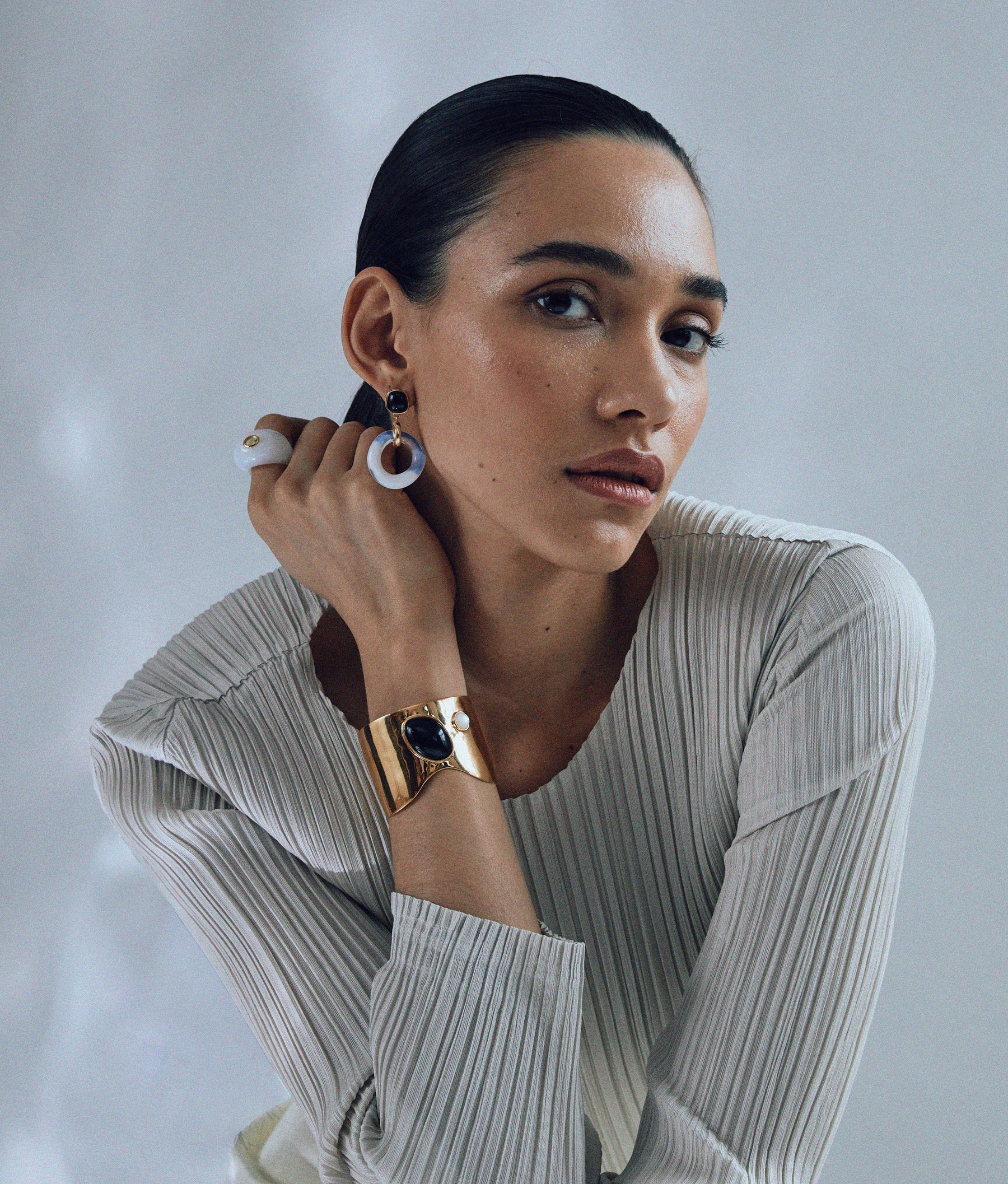 A person with slicked-back hair wears a pleated top, large earrings, and a gold bracelet, elegantly posing with their hand near their face against a neutral background. Their fingers are adorned with vibrant Lizzie Fortunato Monument Rings, each piece a wearable art masterpiece.