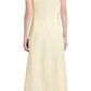 A woman in the Proenza Schouler Candice Dress, a long, cream-colored linen viscose dress with thin straps, viewed from the back. She has wavy hair and is wearing black shoes.