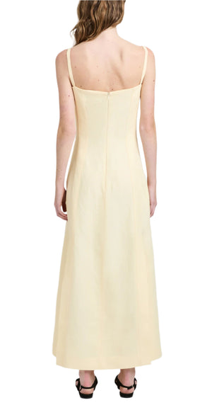 A woman in the Proenza Schouler Candice Dress, a long, cream-colored linen viscose dress with thin straps, viewed from the back. She has wavy hair and is wearing black shoes.